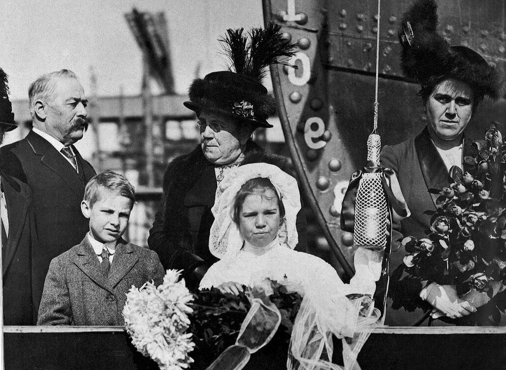 A young girl dressed in white stands ready to break a bottle of champagne hanging in a netted sack as dressed up onlookers wait for the moment.